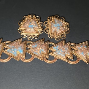 May include: A gold-tone metal jewelry set consisting of a bracelet and a pair of clip-on earrings. Each piece features triangular motifs with a textured, speckled finish in shades of light blue and copper, framed by intricate, decorative filigree borders.