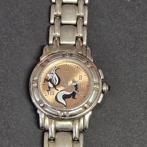 Pu&ograve; includere: Orologio da polso argentato con quadrante marrone chiaro, numeri romani e due moffette dei cartoni animati. L'orologio ha un cinturino argentato e una corona nera. Un accessorio classico.
