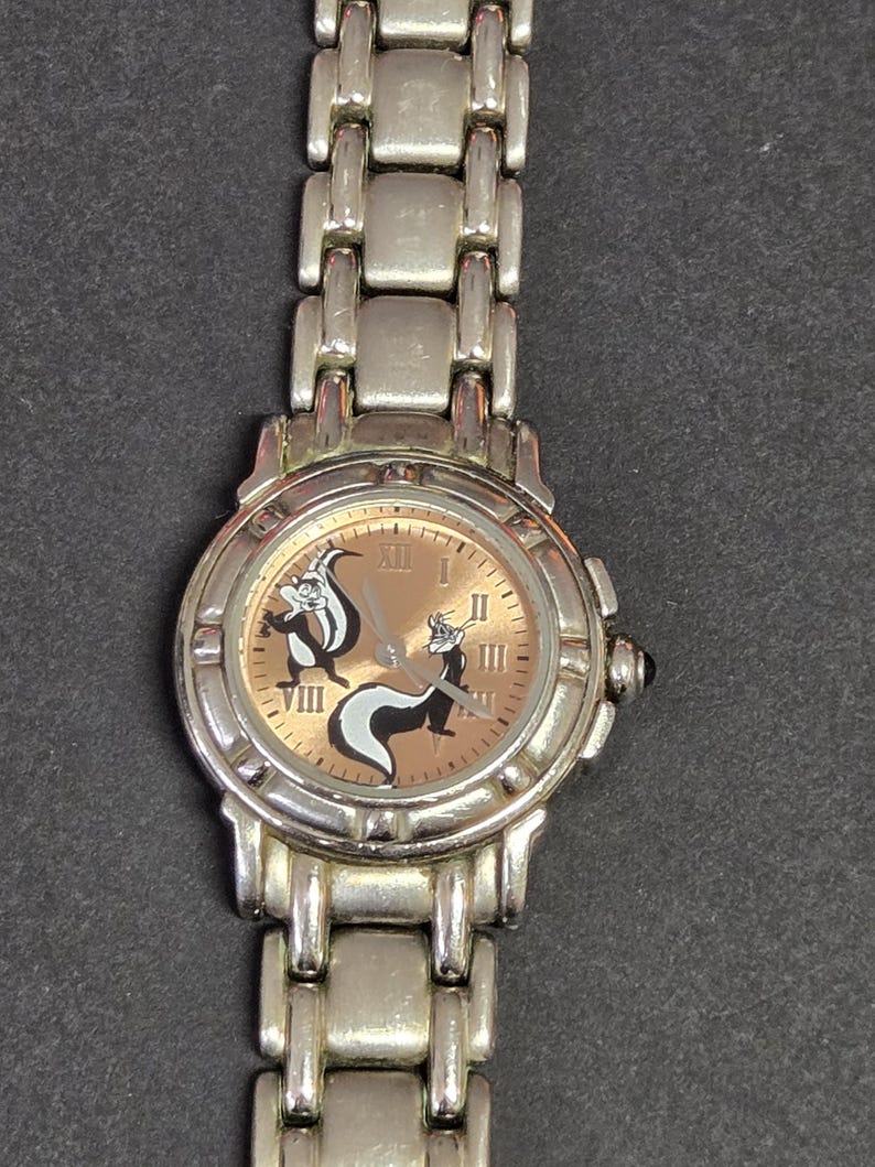 Pu&ograve; includere: Orologio da polso argentato con quadrante marrone chiaro, numeri romani e due moffette dei cartoni animati. L'orologio ha un cinturino argentato e una corona nera. Un accessorio classico.