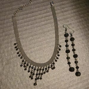 May include: A jewelry set consisting of a choker necklace and a pair of matching drop earrings. The necklace features a metallic mesh band with dangling black faceted beads. The long earrings are composed of a series of black beads on metal links.