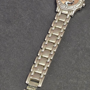 Pu&ograve; includere: Orologio da polso color argento con un personaggio di puzzola dei cartoni animati sul quadrante. L'orologio ha un quadrante rotondo con sfondo marrone chiaro e lancette e indici argentati. Il cinturino &egrave; in metallo argentato.