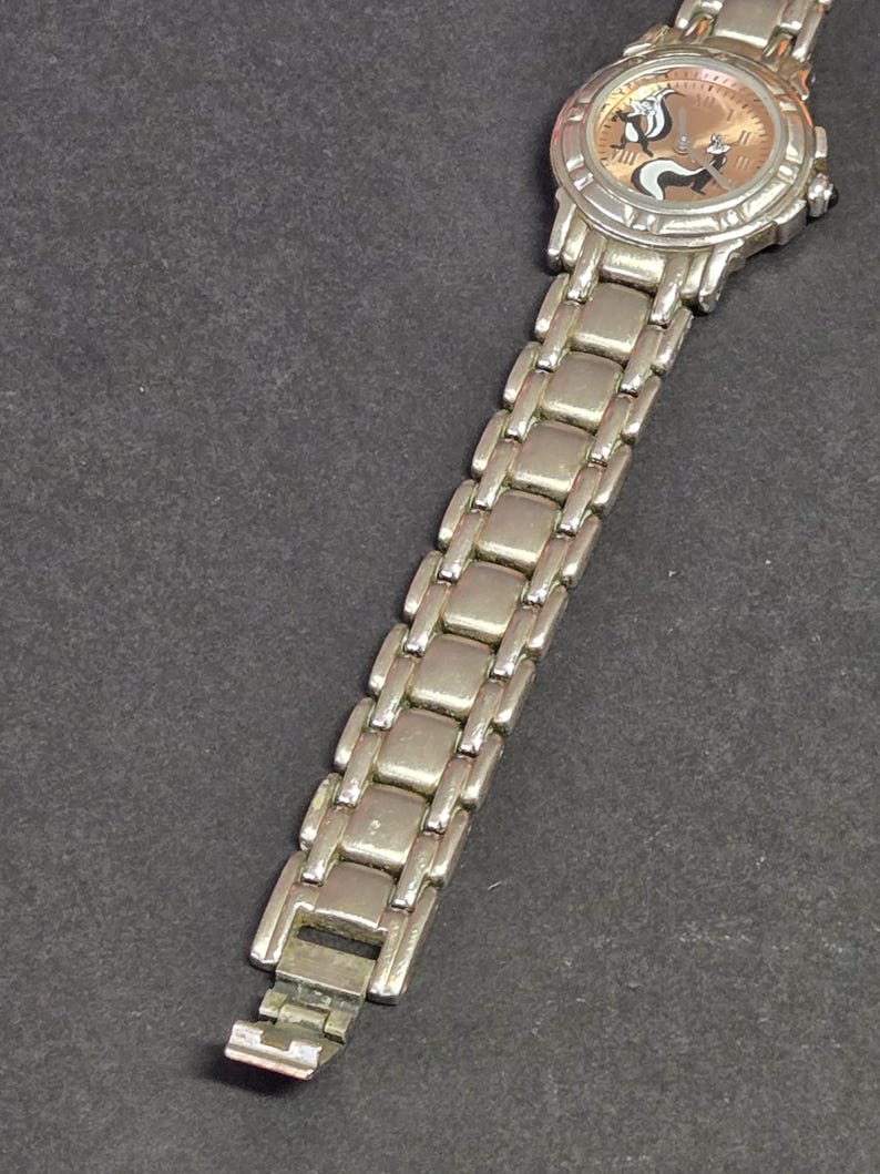 Pu&ograve; includere: Orologio da polso color argento con un personaggio di puzzola dei cartoni animati sul quadrante. L'orologio ha un quadrante rotondo con sfondo marrone chiaro e lancette e indici argentati. Il cinturino &egrave; in metallo argentato.