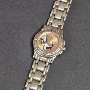 Pu&ograve; includere: Orologio da polso argentato con quadrante marrone chiaro con un design di puzzola dei cartoni animati. L'orologio ha un cinturino argentato con maglie rettangolari. Il quadrante mostra numeri romani e lancette nere.
