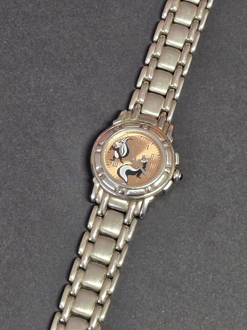 Pu&ograve; includere: Orologio da polso argentato con quadrante marrone chiaro con un design di puzzola dei cartoni animati. L'orologio ha un cinturino argentato con maglie rettangolari. Il quadrante mostra numeri romani e lancette nere.