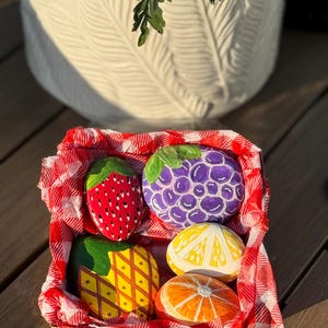 May include: A square cardboard basket lined with red and white checkered tissue paper containing four hand-painted rocks shaped like fruit. The collection includes a red strawberry, a purple cluster of grapes, a yellow pineapple, and citrus slices.