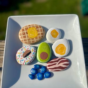 May include: A white square plate displays painted rocks resembling breakfast foods. The rocks include a waffle with butter, two eggs, a donut with sprinkles, an avocado, bacon, and blueberries. The rocks are painted with bright colors.