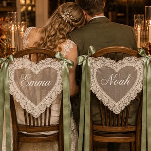 Puede incluir: Dos sillas de madera con fundas de encaje blanco en forma de corazón y lazos de cinta verde salvia. La silla izquierda muestra el nombre Emma en letra cursiva blanca y la derecha el nombre Noah en letra cursiva blanca, centrados en el encaje.