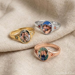 May include: Three personalized signet rings in gold, silver, and rose gold. Each ring features an oval photo and decorative floral accents. The rings are displayed on a neutral fabric background.