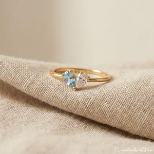 Puede incluir: Un anillo de oro con dos piedras preciosas en forma de pera, una azul claro y otra transparente. El anillo está sobre una tela de color neutro. El nombre de la marca "CrumbleAndGlow" es visible.