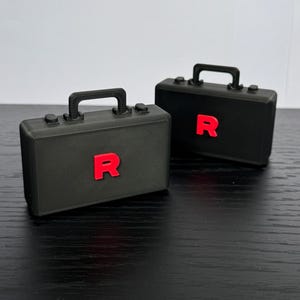 Puede incluir: Dos maletines negros con una letra "R" roja en la parte delantera. Cada maletín tiene un asa negra y dos pequeños botones negros. Los maletines están sobre una superficie oscura.