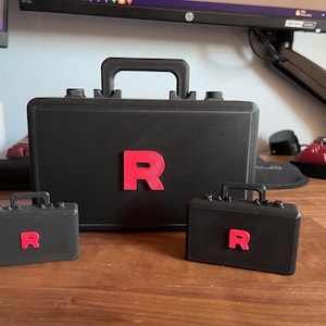 Op de afbeelding: Een set van drie zwarte aktetassen met een rode "R" op elk. De grootste aktetas heeft een handvat aan de bovenkant en de kleinere hebben ook handvatten. De aktetassen staan op een houten oppervlak.