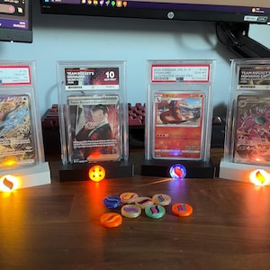 May include: Four graded Pokémon cards in protective cases, displayed on illuminated stands. The cards feature various characters and are accompanied by colourful game tokens. The text on the cards is visible.