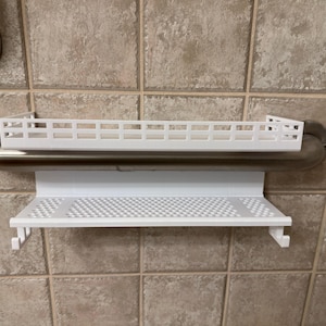 May include: A white, 3D-printed shower shelf with a rectangular design. The shelf has a perforated bottom and is mounted on a metal grab bar. The shelf has two hooks on the bottom for hanging items.