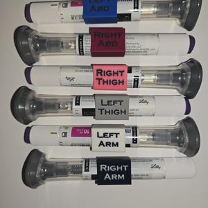 May include: A vertical shot shows six white and clear medical devices with gray caps and purple ends. Each device has a colored label indicating injection site: blue "LEFT ABD", red "RIGHT ABD", pink "RIGHT THIGH", gray "LEFT THIGH", white "LEFT ARM", and black "RIGHT ARM".