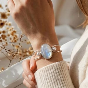 May include: A silver cuff bracelet featuring a large, oval-shaped iridescent moonstone at the center, flanked by two small, round white pearls. The metal band has a polished finish and an open design that wraps around the wrist.