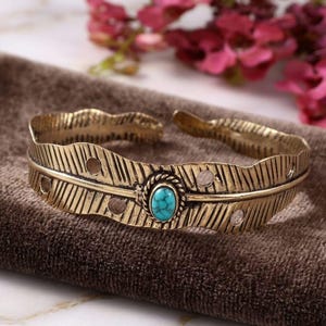 May include: A gold-toned metal cuff bracelet shaped like a feather with a textured surface and small circular cutouts. The centre features an oval-shaped turquoise stone set within a twisted metal border, creating a detailed focal point.