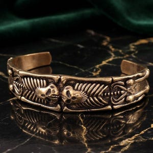 May include: A brass-toned metal cuff bracelet featuring a repeating relief pattern of human skulls and rib cages. The design is segmented into rectangular panels with a weathered, antique finish and an open back for adjustable wear.
