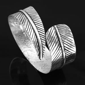 May include: A silver-toned metal cuff bracelet featuring a wrap-around design shaped like two overlapping feathers. The surface of the bracelet displays detailed etched lines mimicking the texture of a feather quill, with a polished finish.