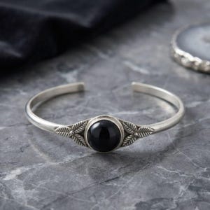 Op de afbeelding: Een zilverkleurige open armband met een centrale ronde zwarte steen. De zetting is aan beide zijden van de steen versierd met gedetailleerde bladmotieven die langs de band doorlopen. Het metaal heeft een gladde en gepolijste afwerking.