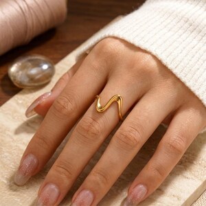 Puede incluir: Un anillo de tono dorado que presenta un diseño de banda continua, lisa y ondulada o serpentina. El anillo tiene una superficie pulida y reflectante, y se lleva en el dedo medio, mostrando una forma fluida y curva alrededor del dedo.