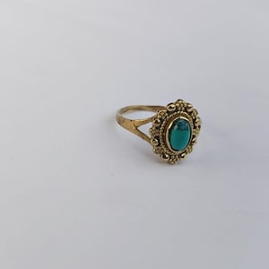 May include: A vintage-style ring with a gold-colored band and setting. The ring features an oval, turquoise-colored stone at the center, surrounded by a detailed, beaded design. The ring is set against a white background.