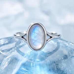 May include: A silver ring featuring a central oval-shaped moonstone with a blue iridescent sheen. The band has a split shank design that connects to the bezel setting, showcasing a polished metallic finish and a smooth, reflective surface.