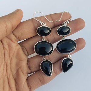 May include: A pair of drop earrings featuring three polished black stones set in silver-toned metal. Each earring consists of a small oval stone at the top, a larger oval stone in the centre, and a teardrop-shaped stone at the bottom.