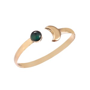 May include: A gold-toned open cuff bracelet featuring a round, dark green cabochon stone on one end and a crescent moon shape on the other. The band is smooth and polished, creating a continuous circular form with an opening between the two ends.
