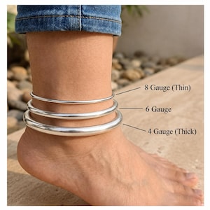 May include: Three smooth, polished silver-tone anklets of varying thicknesses. The top anklet is labeled 8 Gauge (Thin), the middle is 6 Gauge, and the bottom is 4 Gauge (Thick). Each band is a simple, continuous circular loop of metal.