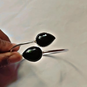 Puede incluir: Una pulsera de puño plateada con dos piedras negras pulidas en forma de lágrima. Las piedras están engastadas en un bisel y están conectadas por una banda delgada y abierta. La pulsera se muestra sobre un fondo blanco.