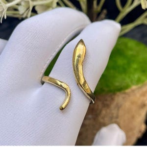 Könnte beinhalten: Ein goldfarbener offener Ring mit einem glatten, geschwungenen Design, das sich teilweise um den Finger legt. Die Metalloberfläche hat ein poliertes Finish mit leicht zulaufenden Enden, was eine schlichte und durchgehende Form erzeugt.