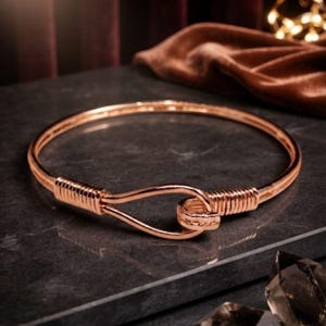 Puede incluir: Un brazalete rígido de metal en tono oro rosa con un cierre de tipo gancho. La banda circular presenta detalles decorativos de alambre enrollado a ambos lados del mecanismo de cierre, creando un acabado metálico pulido y uniforme.