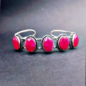 Könnte beinhalten: Ein silberfarbenes Armreif-Armband mit fünf ovalen, leuchtend roten Edelsteinen. Jeder Edelstein ist in einen perlenbesetzten Rahmen gefasst. Das Armband hat ein offenes Design, die Enden sind nach außen gebogen.