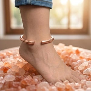May include: A smooth, polished copper anklet with an open-ended design. The metallic band features a rounded, cylindrical profile and a warm, reddish-brown hue, encircling the ankle above a bed of textured, translucent pink salt crystals.