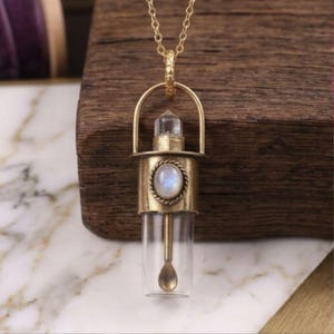 Puede incluir: Un collar con colgante de latón que presenta un vial de vidrio transparente con una pequeña cuchara de latón en su interior. El vial está rematado con una tapa de latón, una punta de cristal transparente y un cabujón de piedra lunar.