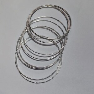 Puede incluir: Una colección de pulseras finas de metal plateado. Las pulseras son circulares y parecen estar hechas de un material brillante y reflectante. Están apiladas, creando un efecto de capas.