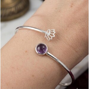 May include: A silver open cuff bracelet featuring a polished round purple amethyst gemstone on one end and a cutout lotus flower design on the other end. The metal band is smooth and circular, creating a bypass style that remains open at the centre.