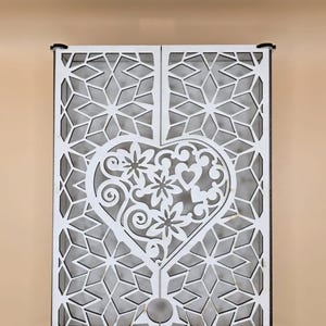 May include: A white wooden wall decor piece with a heart and floral design. The heart is the focal point, filled with intricate floral and swirl patterns. The background features a geometric starburst pattern. The piece is framed in black.