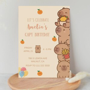Może przedstawiać: Zaproszenie urodzinowe z jasnobeżowym tłem. Zaproszenie zawiera tekst "Let's Celebrate Amelia's Capy Birthday" oraz ilustracje kapibar i pomarańczy. Uwzględniono r&oacute;wnież datę, godzinę i miejsce.