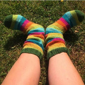 May include: A pair of hand-knitted socks with horizontal stripes in green, blue, orange, and pink. The socks are worn on legs resting on green grass. The cuffs and toes are a solid green colour.