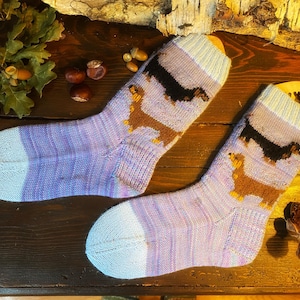 May include: A pair of knitted socks in shades of lavender, blue, and white, featuring dachshund dog designs in brown and black. The socks are displayed on a wooden surface, showcasing a cozy, handmade aesthetic.