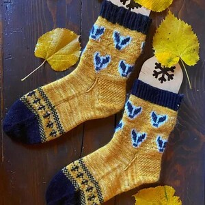 Pode incluir: Um par de meias feitas à mão em amarelo mostarda com detalhes em azul marinho. As meias apresentam um padrão repetido de formas azuis e punhos e dedos dos pés em azul marinho. Folhas de outono amarelas acompanham as meias. As meias têm cerca de 25 cm de comprimento.