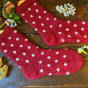 May include: A pair of red knit socks with white polka dots. The socks are displayed on a wooden surface, with flowers in the background. The socks are a cozy and warm accessory.