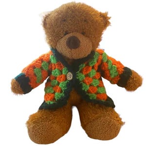 Könnte beinhalten: Ein brauner Teddybär mit einer bunten gehäkelten Jacke. Die Jacke hat ein Muster aus orangefarbenen und grünen Quadraten mit dunkelgrüner Bordüre und einem einzelnen Knopfverschluss. Der Bär hat eine weiche, plüschige Textur.