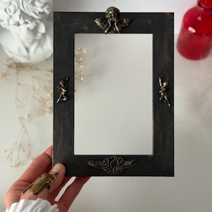 May include: A rectangular black framed mirror with bronze cherub and scroll decorations. The mirror is held by a hand, and the frame has a distressed finish. The overall theme is vintage decor.
