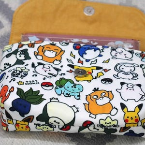 Könnte beinhalten: Weiße Stofftasche mit buntem Cartoon-Charakter-Druck. Die Tasche hat einen Druckknopfverschluss und ein gelbes Innenfutter. Der Druck enthält verschiedene Charaktere, Pokebälle und den Text "キキララ DOODLES".