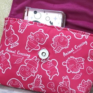May include: A pink pouch with a white cartoon character pattern and the text "Animal Crossing MOVE OUT". The pouch has a magnetic snap closure and a magenta interior. A clear phone case is visible inside.