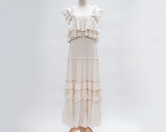 Puede incluir: Un vestido largo crema con volantes sobre un maniquí de madera. El vestido presenta un escote cuadrado, mangas con volantes y múltiples capas de tela. El vestido es largo hasta el suelo y tiene un estilo bohemio fluido.