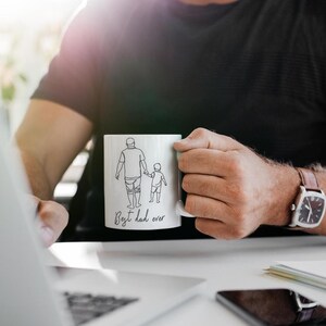May include: White ceramic mug with a black line drawing of a parent and child holding hands. The text "Best dad ever" is written below the illustration. The mug is held by a person wearing a black shirt.