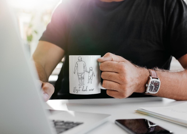May include: White ceramic mug with a black line drawing of a parent and child holding hands. The text "Best dad ever" is written below the illustration. The mug is held by a person wearing a black shirt.